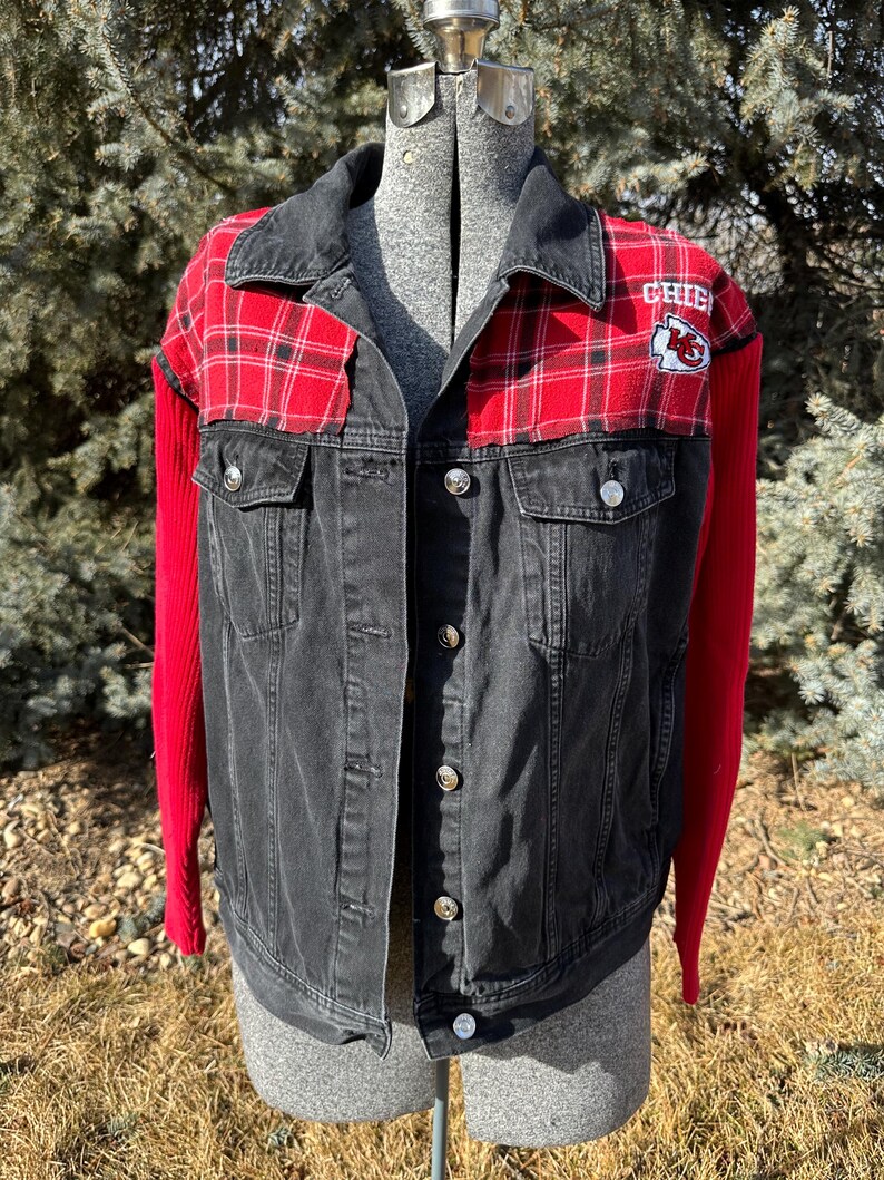 Puede incluir: Chaqueta vaquera negra con detalles de cuadros rojos en los hombros y la parte superior del pecho. Las mangas son de tejido de punto rojo. La chaqueta tiene botones plateados y un parche con la palabra "CHIEFS" y un logotipo.