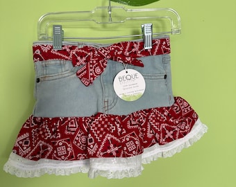 May include: A light blue denim skirt featuring a red and white bandana print waistband with a tied bow, a matching red and white bandana print ruffled hem, and a white eyelet lace trim. A circular tag with the text BEQUE is attached.