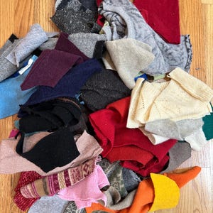May include: A collection of assorted fabric scraps in various colors and textures, including red, orange, yellow, pink, blue, grey, black, and cream pieces. The pile features a mix of solid, patterned, and knit materials of different shapes.