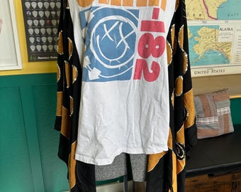 May include: A white t-shirt with the text "blink-182" in orange and pink, featuring the band's logo. The shirt is paired with a black and gold patterned shawl with fringe.