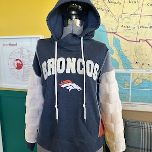 May include: A dark blue hooded sweatshirt featuring the word BRONCOS in white textured lettering across the chest, with a white horse head logo below. The long sleeves are made of a white, quilted, fuzzy material, and the hood has white drawstrings.