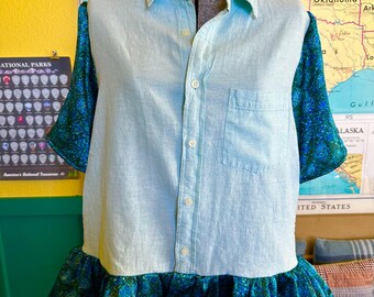 May include: A light blue button-down shirt with short sleeves and a ruffled hem. The sleeves and hem are made of a patterned teal fabric. The shirt has a collared neckline and a single chest pocket.
