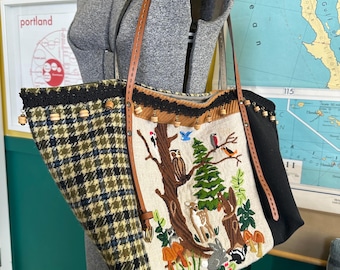 May include: A large tote bag with a woodland scene embroidered on the front. The bag features a black and green plaid pattern on one side, a black panel on the other, and brown leather straps. The embroidered scene includes trees, animals, and mushrooms.