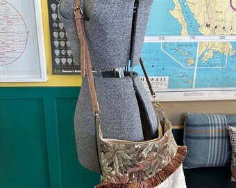 May include: A crossbody bag with a tapestry design featuring leaves in shades of green, brown, and cream. The bag has a cream-colored bottom with an embroidered pattern and a brown fringe. It has a brown leather strap.