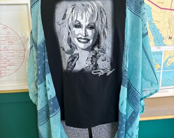 May include: Black t-shirt featuring a grayscale portrait of a smiling woman with the signature "Dolly". A sheer, light blue paisley patterned kimono is layered over the shirt, creating a unique and stylish look.