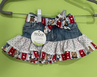 May include: A denim skirt with a red and white patterned ruffle and bow. The bow and trim feature teddy bears, hearts, and floral designs. The skirt is hanging on a gray hanger.