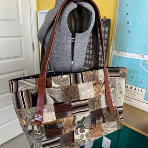 May include: A large tote bag featuring a patchwork design of various woven fabrics in shades of brown, tan, black, and cream. The bag has two dark brown braided leather shoulder straps attached with brass-toned buckles and metal rivets.