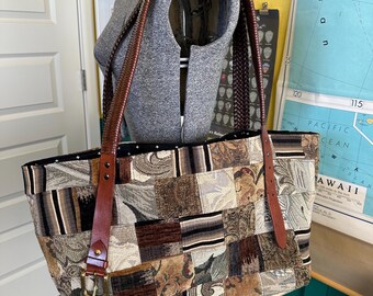 May include: A large tote bag featuring a patchwork design of various woven fabrics in shades of brown, tan, black, and cream. The bag has two dark brown braided leather shoulder straps attached with brass-toned buckles and metal rivets.