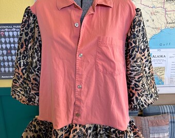 May include: A coral-colored button-down shirt with leopard print sleeves and a ruffled hem. The shirt is displayed on a mannequin. The sleeves and hem feature a brown, black, and tan animal print pattern. The shirt has a single chest pocket.