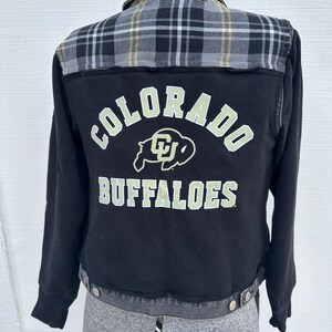 May include: A black jacket featuring a grey, black, and yellow plaid yoke and collar. The back displays the text COLORADO BUFFALOES in white lettering with a central buffalo logo. The jacket has long sleeves, a button-front closure, and a hem.