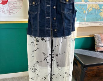 May include: A sleeveless denim vest with a dark wash, featuring button closures and two chest pockets. The vest is paired with a sheer, white, floor-length skirt adorned with a black floral pattern. The garment is displayed on a mannequin.