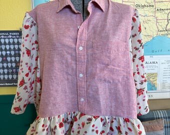 May include: A pink button-down shirt with floral patterned sleeves and a ruffled hem. The sleeves are sheer with a cream background and red floral print. The shirt is displayed on a mannequin in front of a map.