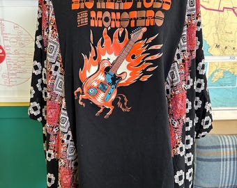 May include: Black t-shirt with the text "BIG HEAD TODD AND THE MONSTERS" in orange and brown. The shirt features a graphic of a guitar engulfed in flames. The sleeves are patterned with red, black, and white geometric designs.
