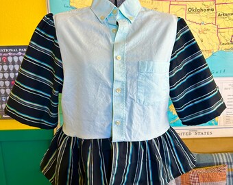May include: A light blue button-down shirt with striped sleeves and a ruffled hem. The sleeves feature black and blue stripes. The shirt is displayed on a mannequin.