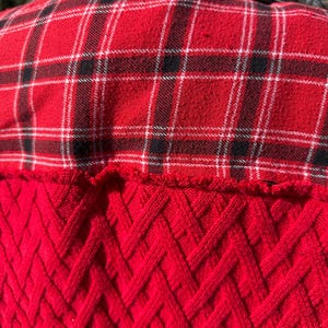 Puede incluir: Una camisa de franela a cuadros rojos y negros con cuello de mezclilla negro. La parte inferior de la espalda de la camisa presenta un dise&ntilde;o de punto de cable rojo texturizado. La camisa es una mezcla de texturas y patrones.