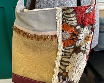 May include: A patchwork shoulder bag with a variety of textures and colors. The bag features a mustard yellow quilted fabric, a section with floral embroidery in red, orange, and cream, and a gray top. The bag has a burgundy bottom and is trimmed with cream-colored lace and brown beads.