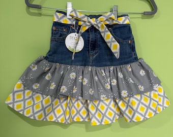 May include: A denim skirt with a gray and white daisy print ruffle and a yellow and gray diamond pattern. The skirt has a matching bow and a tag that reads "BEQ E".