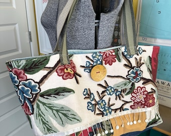 May include: A large tote bag with a floral embroidered design in red, blue, and green on an off-white background. The bag has a wooden button closure and a fringe detail along the bottom. The straps are olive green and gray.