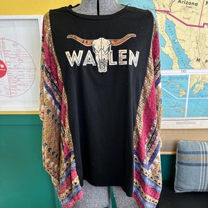 May include: A black short-sleeve top featuring a graphic of a longhorn skull above the text "WAYLEN" in white. The sides of the top are made of a multi-colored fabric with geometric patterns in shades of mustard yellow, magenta, and blue.