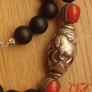 May include: A black onyx beaded necklace with a silver pendant featuring a stylized animal design. The pendant is flanked by two red coral beads.