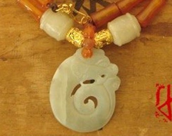 Op de afbeelding: Een ketting met een lichtgroene hanger en oranje en lichtgroene kralen. De hanger heeft een gesneden ontwerp. De ketting ligt op een houten oppervlak met Aziatische karakters.