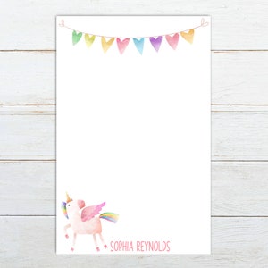 May include: White stationery with a watercolor illustration of a pink unicorn with a rainbow mane and tail. A banner of colorful pennants hangs at the top. The name "SOPHIA REYNOLDS" is printed in pink at the bottom.