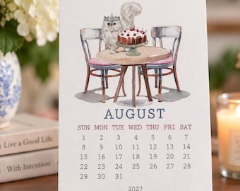 May include: A white desk calendar page for August 2027 featuring a watercolor illustration of a fluffy white cat wearing a crown, standing at a small round table with a chocolate cake topped with cherries, flanked by two wooden chairs.