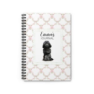 May include: Spiral-bound journal with a white cover featuring a repeating pattern of pink bows and green leaves. The center has a square frame with the text "Emma's JOURNAL" and a watercolor illustration of a black dog.