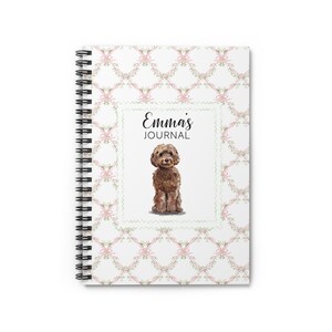 May include: Spiral-bound journal with a white cover featuring a repeating pattern of pink bows and green leaves. The center has a watercolor illustration of a brown dog and the text "Emma's JOURNAL".