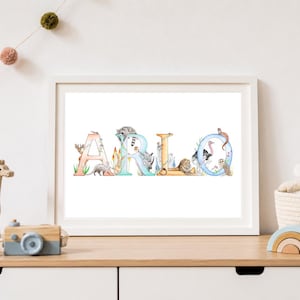 Op de afbeelding: Een ingelijste muurkunstprint met de naam ARLO in grote, kleurrijke letters. Elke letter is versierd met gedetailleerde aquarelillustraties van diverse wilde dieren, waaronder een wasbeer, leeuw, struisvogel en uil, op een witte achtergrond.
