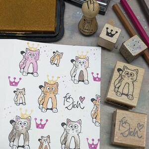 May include: A white card with a repeating pattern of hand-drawn cats wearing crowns. The card has the words "f&uuml;r dich" written on it in cursive. There are several wooden stamps with cat and crown designs, as well as a stamp that says "f&uuml;r dich".