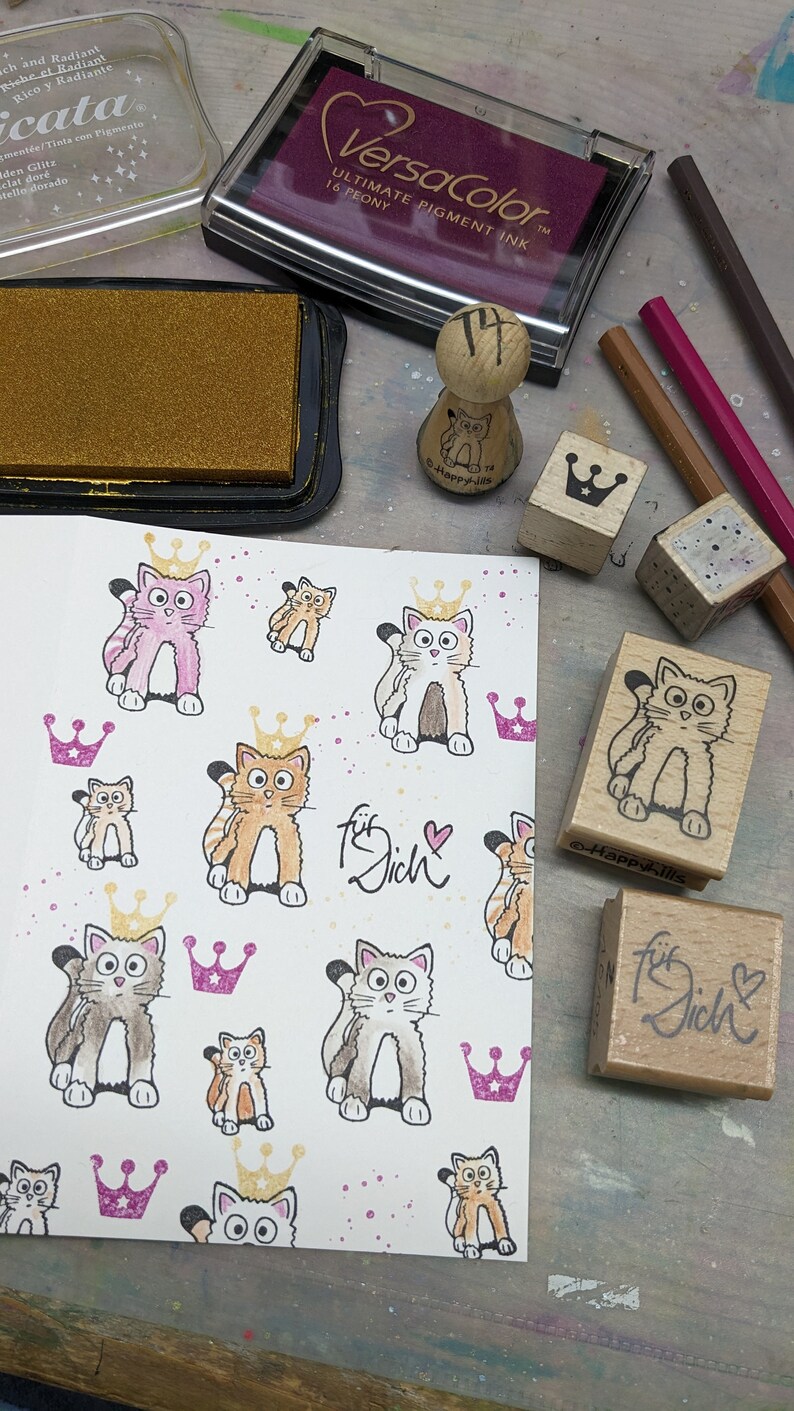 May include: A white card with a repeating pattern of hand-drawn cats wearing crowns. The card has the words "f&uuml;r dich" written on it in cursive. There are several wooden stamps with cat and crown designs, as well as a stamp that says "f&uuml;r dich".