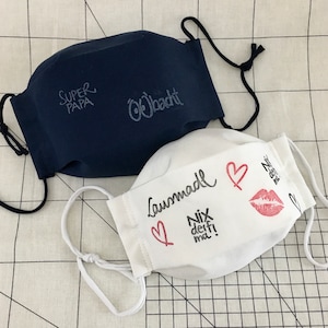 May include: Two fabric face masks. One is navy blue with the text "Super Papa" and "Obacht" in white. The other is white with the text "Lausmadel" and "Nix deif! ma!" in black, a red heart, and a red lipstick print.