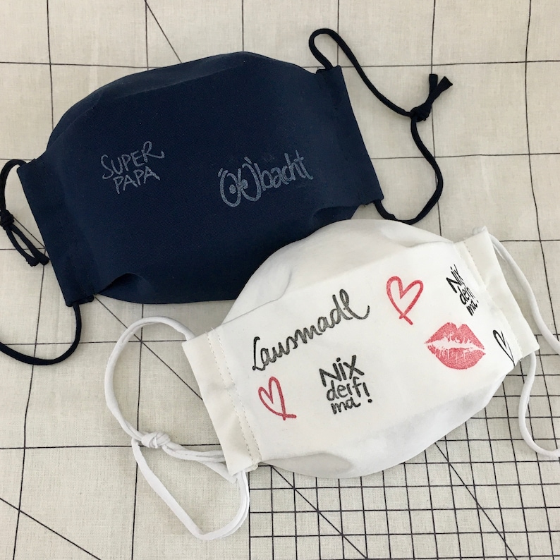 May include: Two fabric face masks. One is navy blue with the text "Super Papa" and "Obacht" in white. The other is white with the text "Lausmadel" and "Nix deif! ma!" in black, a red heart, and a red lipstick print.