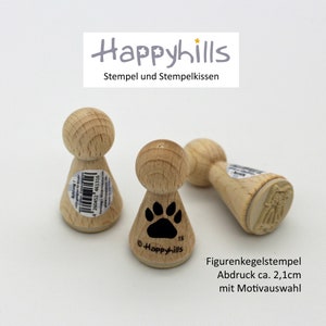 May include: Three wooden cone-shaped stamps with different designs. The stamp on the left has a barcode and text. The middle stamp has a black paw print. The stamp on the right has a white angel design. The stamps are labeled "Happyhills" and "Figurenkegelstempel".