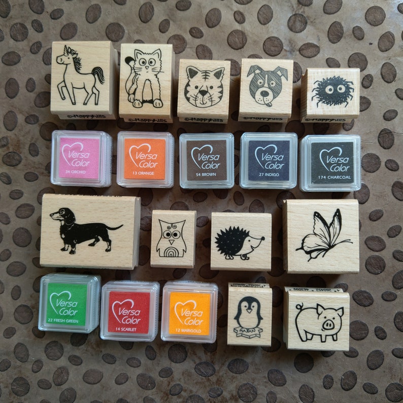 May include: A collection of 16 wooden stamps featuring various animal illustrations, including a unicorn, cat, tiger, dog, monster, dachshund, owl, hedgehog, butterfly, penguin, and pig. The stamps are accompanied by 8 ink pads in various colors, including orchid, orange, brown, indigo, charcoal, green, scarlet, and marigold.