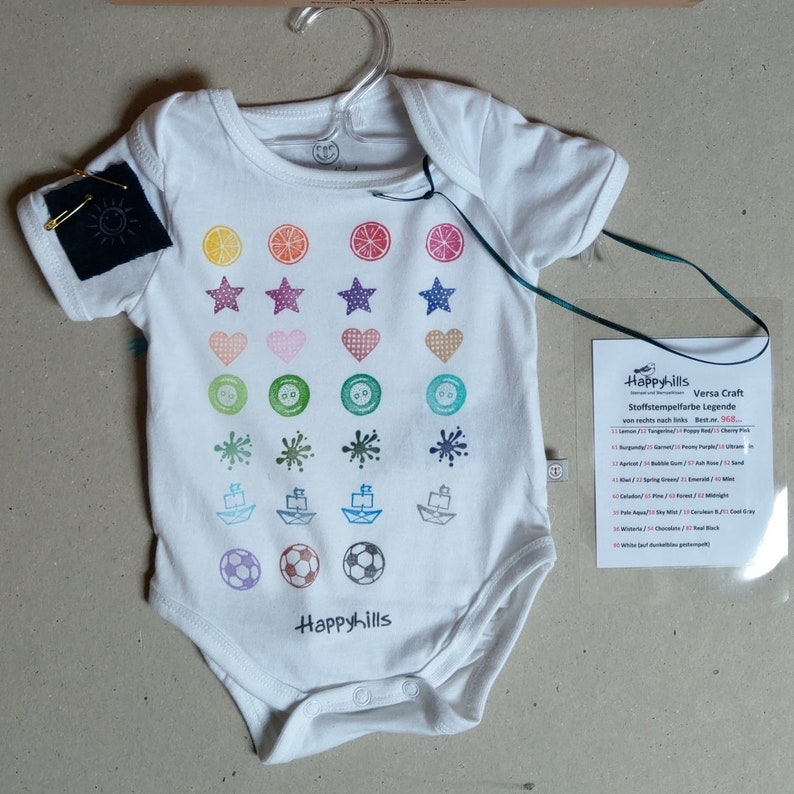 May include: White baby onesie with a colorful pattern of stamped images, including lemons, stars, hearts, buttons, paint splatters, paper boats, and soccer balls. The onesie has the text "Happyhills" printed on the bottom.