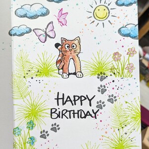 May include: A handmade greeting card with a cartoon cat, butterflies, clouds, a sun, and flowers. The card says "Happy Birthday" in black marker.