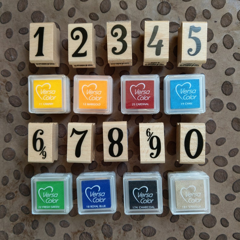 Op de afbeelding: Een set van tien houten cijferstempels met zwarte inkt en een set van acht inktkussens in verschillende kleuren. De inktkussens zijn gelabeld met de merknaam Versa Color en een kleurnaam.