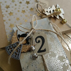Op de afbeelding: Een bruine papieren adventskalender met een zilveren en wit patroon. De kalender heeft een houten engelversiering en een label met het nummer "2" erop.