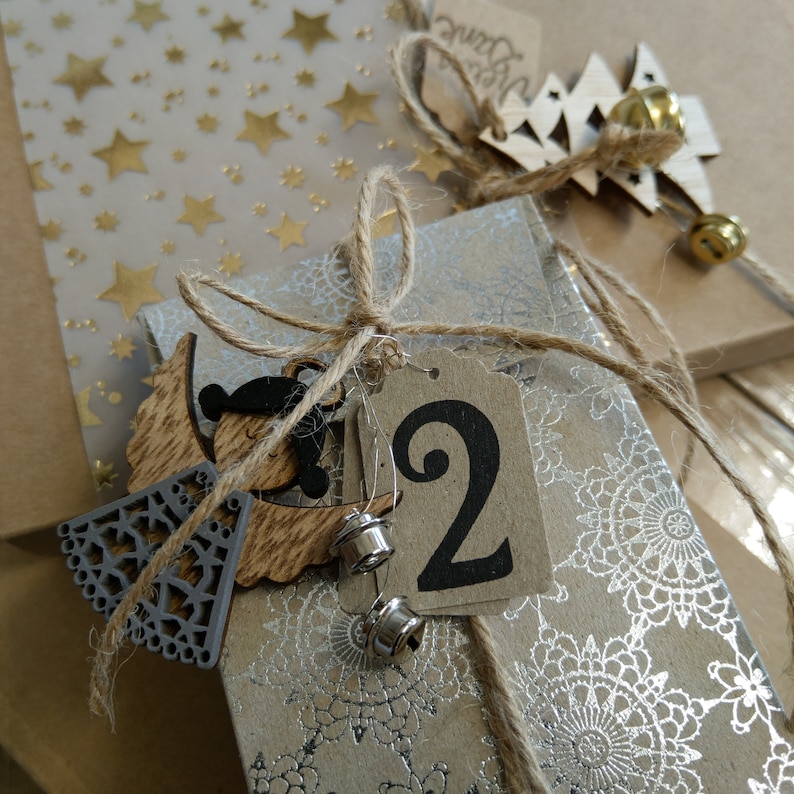 Op de afbeelding: Een bruine papieren adventskalender met een zilveren en wit patroon. De kalender heeft een houten engelversiering en een label met het nummer "2" erop.