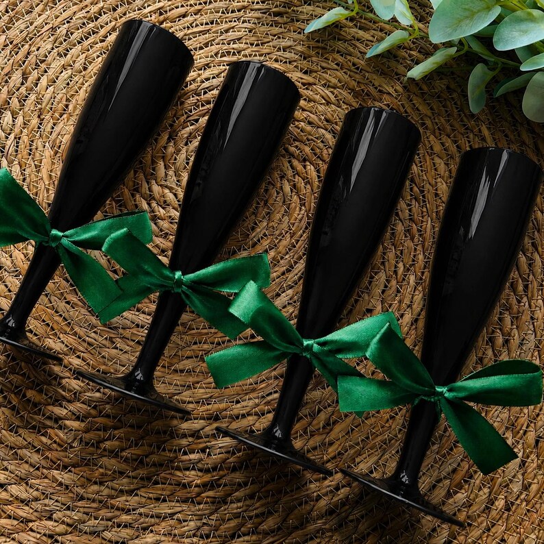 May include: Four tall, slender champagne flutes made of solid black glass. Each flute features a smooth, glossy finish and is tied with a vibrant emerald green satin ribbon bow around the stem, creating a distinct contrast against the dark glass.