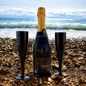May include: A bottle of Chapel Down Brut English sparkling wine with a gold foil neck wrap, positioned between two tall, slender, solid black champagne flutes. The bottle label features the text Chapel Down English Sparkling Wine Brut.