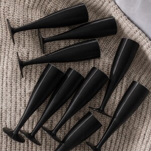 May include: A collection of eight solid black champagne flutes arranged in a scattered pattern. Each flute features a slender, elongated bowl, a thin stem, and a circular base, all finished in a uniform, glossy black color throughout the entire glass.
