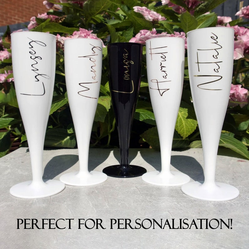 May include: Five champagne flutes, four white and one black, with gold lettering. The flutes are personalised with the names "Lindsay", "Mandy", "Olivia", "Harriet", and "Natalie".