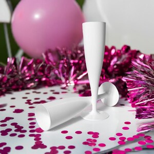 May include: Two white plastic champagne flutes on a white surface with pink confetti. The flutes are stacked on top of each other.