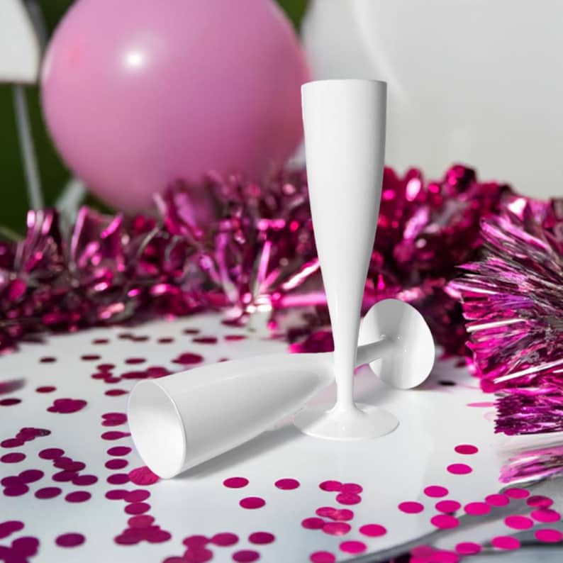 May include: Two white plastic champagne flutes on a white surface with pink confetti. The flutes are stacked on top of each other.