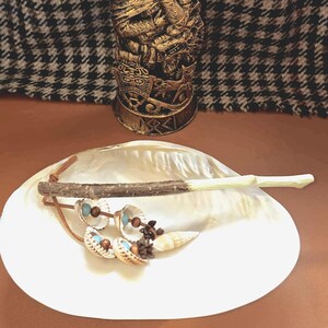 Peut inclure: Une statue dorée sculptée d'une personne vêtue d'un vêtement drapé, sur une base aux motifs complexes. Un grand coquillage blanc contient une baguette en bois, des boucles d'oreilles et de petits objets décoratifs. Le fond est un motif pied-de-poule noir et blanc.