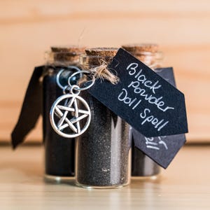 May include: Three small glass bottles filled with black powder, labeled "Black Powder Doll Spell". Each bottle has a silver pentagram charm attached.