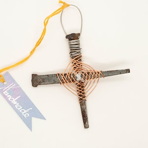 Puede incluir: Adorno de cruz hecho a mano con clavos oxidados y alambre de cobre. La cruz está atada a una cuerda amarilla con una etiqueta que dice "Hecho a mano".